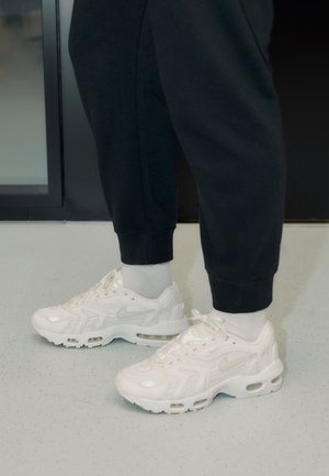 Personne portant des baskets blanches Nike, des chaussettes blanches et un pantalon noir, debout sur un sol clair tacheté près d'une porte en verre.
