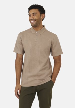 Uomo che sorride, indossa una polo beige a maniche corte con motivi a diamante e trecce, pantaloni verde oliva, con una mano in tasca.