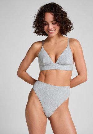 Femme aux cheveux bouclés souriante, portant un slip taille haute gris et un bralette triangle assorti, debout devant un fond uni.
