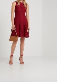 Femme portant une robe rouge en dentelle sans manches et des sandales à talons hauts rouges, tenant un petit sac à main marron avec une bandoulière en chaîne dorée.
