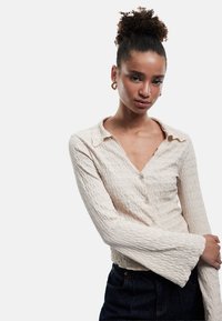 Textureret, lys beige bluse med V-udskæring, lange klokkeærmer og knapper langs fronten. Blødt materiale med et bølget mønster.