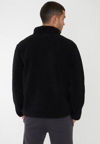 Schwarze Fleecejacke mit hohem Kragen, oversized Schnitt, strukturierte Oberfläche und ohne sichtbare Hardware; getragen über grauen Hosen.