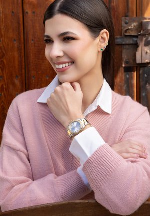 Orologio dorato e argentato con quadrante blu sul polso. Maglione testurizzato rosa chiaro con una camicia bianca visibile sotto.