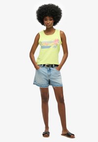Lichtgele tanktop met een afgedrukte strandscène, gecombineerd met versleten denim shorts en zwarte sandalen. Beschikt over een ronde halslijn en een mouwloos ontwerp.