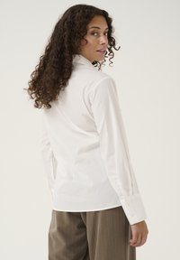 Camicia bianca con colletto, maniche lunghe e finitura liscia, abbinata a pantaloni beige a righe. Vista posteriore mostrata.