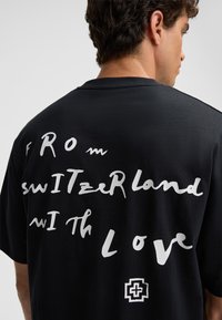 Schwarzes T-Shirt mit weißem handgeschriebenem Text auf dem Rücken: "FROM switZerLand wiTh Love." Enthält ein kleines Logo am unteren Rand.