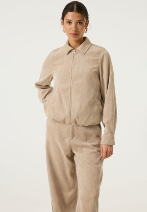 Donna che indossa una giacca beige in velluto a coste con zip e pantaloni abbinati, con orecchini a cerchio dorati, in piedi davanti a uno sfondo chiaro e uniforme.