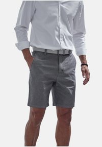 Graue Shorts aus leichtem Stoff, mit flachem Bund, Gürtelschlaufen und seitlichen Taschen. Kombiniert mit einem weißen Langarmshirt.