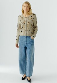 Lichtbeige gebreide cardigan met pailletten en twee voorzakken, gecombineerd met lichtblauwe wijde jeans en zwarte puntige schoenen.
