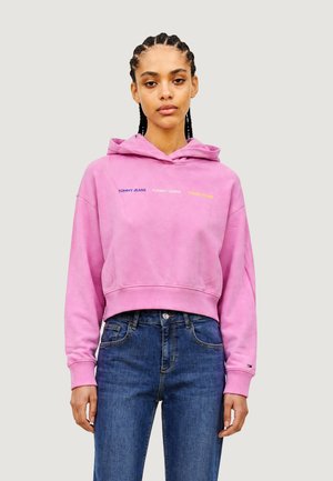 Giovane donna con capelli intrecciati che indossa una felpa corta rosa e jeans blu, in piedi davanti a uno sfondo chiaro e semplice.