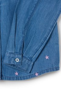 Camisa de mezclilla azul con mangas largas, puños abotonados, con estrellas rosas bordadas en el dobladillo como detalle decorativo.