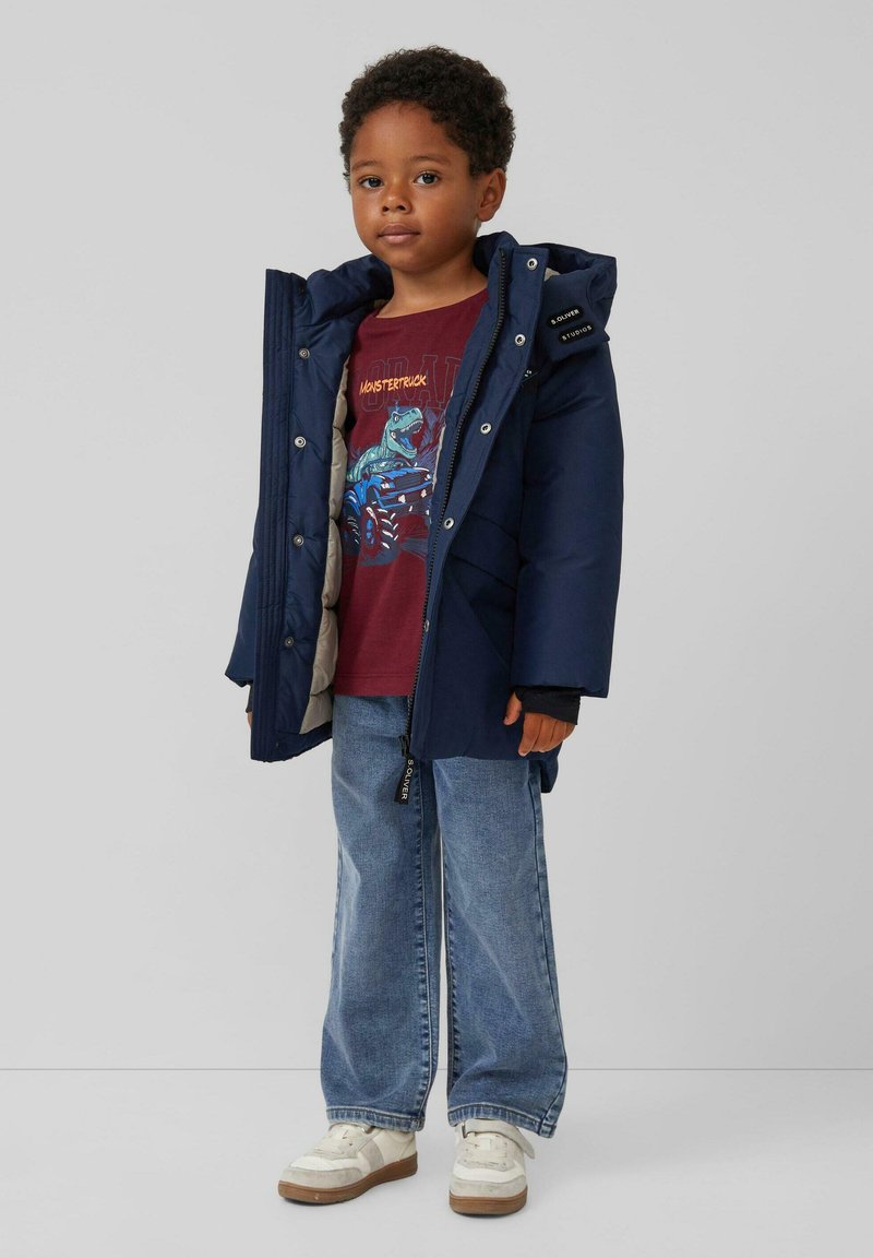 Marineblauwe pufferjack met een capuchon, grijze voering en decoratieve ritsen. Draag het over een bordeauxrood graphic t-shirt en blauwe wijde jeans. Witte sneakers.
