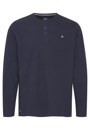 Tamsiai mėlyna ilgomis rankovėmis marškiniai, pagaminti iš medvilnės; turi apvalų iškirpimą, tris sagas ir mažą logotipo siuvinėjimą krūtinėje.