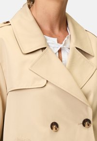 Beige dobbeltknappet frakke med store klapper, fremstillet af glat stof, med mørke knapper og en hvid skjorte med krave nedenunder.