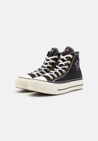 Zapatillas de lona negras de caña alta con cordones blancos, ojales dorados y detalles en rosa y azul en el cuello y en la punta. Suela media blanca gruesa.
