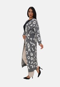 Lungo cardigan grigio con stampa leopardata bianca, tessuto lavorato a maglia, apertura frontale, due tasche e polsini a coste. Abbinato a pantaloni beige e tacchi neri.