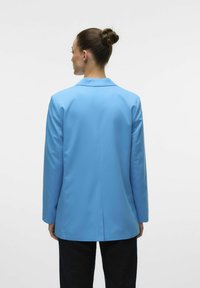 Blazer azzurro chiaro con un design strutturato, maniche lunghe, apertura posteriore singola e una texture di tessuto liscia. Indossato sopra pantaloni scuri.