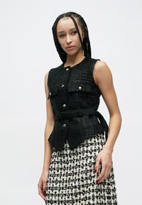 Schwarze Tweedweste mit Fronttaschen, silbernen Knöpfen und tailliertem Gürtel. Kombiniert mit einem karierten schwarz-weißen Rock aus strukturierter Stoffqualität.