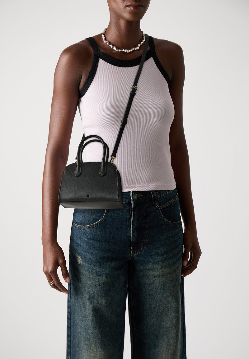 Borsa a mano in pelle nera con forma strutturata, manici doppi e tracolla staccabile. Indossata sopra una canotta grigia chiara e jeans larghi blu.