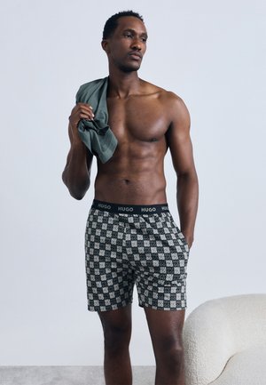 Moški brez srajce stoji z roko v žepu, drži sivo srajco čez ramo in nosi črno-bele vzorčaste HUGO boksarice.