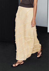 Jupe maxi beige en couches avec des panneaux texturés et ondulés et des détails à franges. Associée à des sandales plates brunes et un haut sombre.