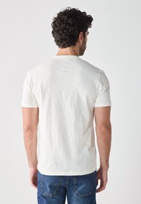 Camiseta blanca de manga corta hecha de un tejido ligero y texturizado. Presenta un escote redondo y un diseño ajustado, visible en la parte posterior.