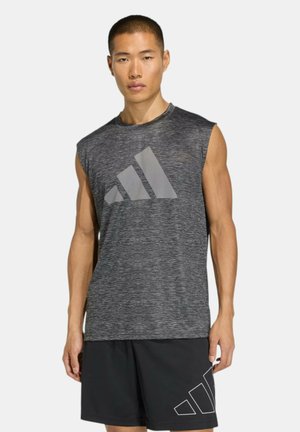 Giovane uomo che indossa una canotta grigia Adidas e pantaloncini neri Adidas, in piedi davanti a uno sfondo chiaro e uniforme.