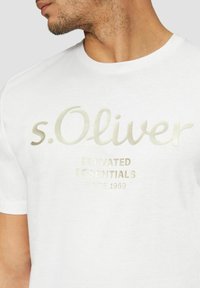 White cotton t-shirt featuring gold foil printed brand name and text "ELEVATED ESSENTIALS SINCE 1969." Classic crew neckline, short sleeves.