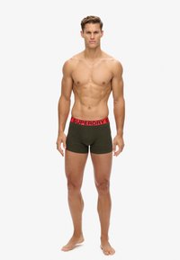 Grüne Boxershorts mit einem roten, gebrandeten Bund, die über eine weiche Textur und eine körpernahe Passform verfügen. Das Modell steht.