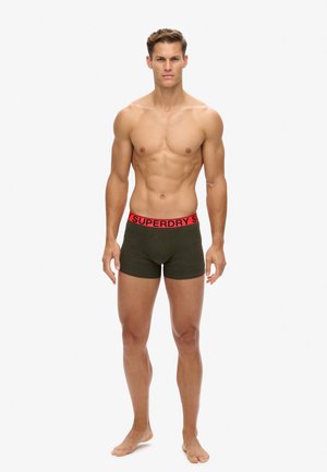 Grüne Boxershorts mit einem roten, gebrandeten Bund, die über eine weiche Textur und eine körpernahe Passform verfügen. Das Modell steht.