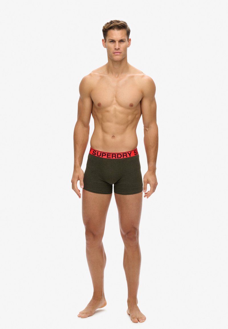 Grüne Boxershorts mit einem roten, gebrandeten Bund, die über eine weiche Textur und eine körpernahe Passform verfügen. Das Modell steht.