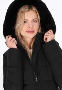 Geacă puffer neagră cu glugă din blană artificială, cu fermoar frontal și design capitonat. Geaca are o textură netedă și este purtată deschisă la guler.