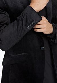 Zwart fluwelen blazer met een satijnglans, voorzien van een enkele zak, vier zwarte knopen op de mouw en een getailleerd kraagontwerp.