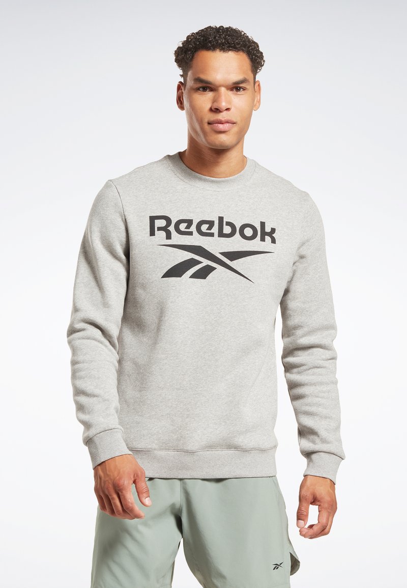 Joven vestido con un sudadera ligera gris de Reebok y pantalones cortos verde salvia, de pie contra un fondo blanco liso.