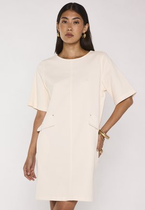 Femme aux longs cheveux foncés porte une robe crème à manches courtes longueur genoux avec poches avant, boucles d'oreilles créoles dorées, bracelets et bagues.
