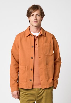Jeune homme aux cheveux châtains porte une veste orange boutonnée sur un polo orange et un pantalon olive, debout devant un fond blanc uni.