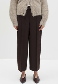 Femme portant un pantalon large marron foncé, un cardigan beige à boutons en peluche et des chaussures pointues marron sur fond blanc.