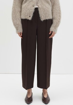 Vrouw in een donkerbruine wijde broek, een beige fuzzy knoopcardigan en puntige bruine schoenen tegen een witte achtergrond.