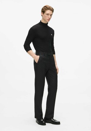 Jeune homme portant un pull à col roulé noir, un pantalon noir à motifs et des chaussures habillées noires, debout avec une main dans la poche.