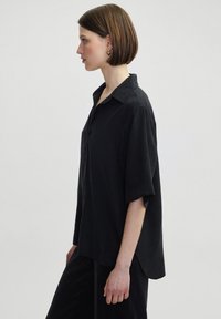 Black short-sleeve shirt features a collared neck, button front, and loose fit. Soft fabric with a smooth texture and gentle drape.