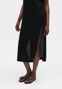 Γυναίκα που φοράει μαύρο πλεκτό αμάνικο φόρεμα με πλευρικές σχισμές, σε συνδυασμό με μαύρα σανδάλια τύπου φλιπ-φλοπ, μπροστά από απλό φόντο.