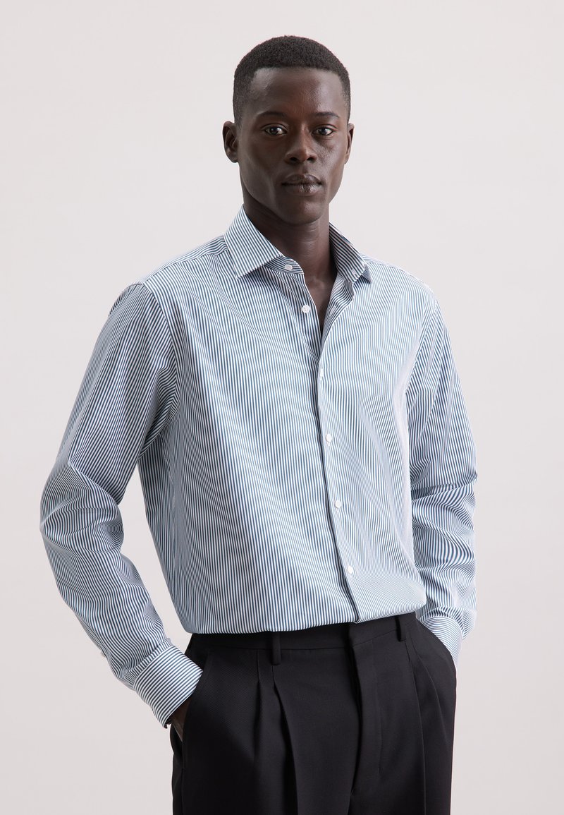 Button-up shirt met verticale blauw-witte strepen, lange mouwen en een klassieke kraag. G gedragen met zwarte broek.