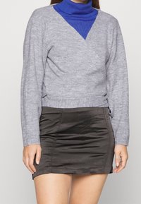 Grå omslagströja med långa ärmar över en blå polotröja, kombinerad med en svart satinminikjol. Mjuk stickad textur, avslappnad design.