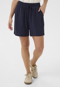 Námornícke ľanové šortky s elastickým pásom, šnúrkou na stiahnutie a bočnými vreckami. V kombinácii so béžovými teniskami s hnedými pruhmi.