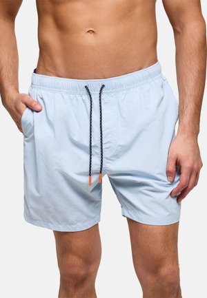 Hombre con bañador azul claro con cordones negros y puntas naranjas, manos en los bolsillos, mostrando la zona media y los muslos.