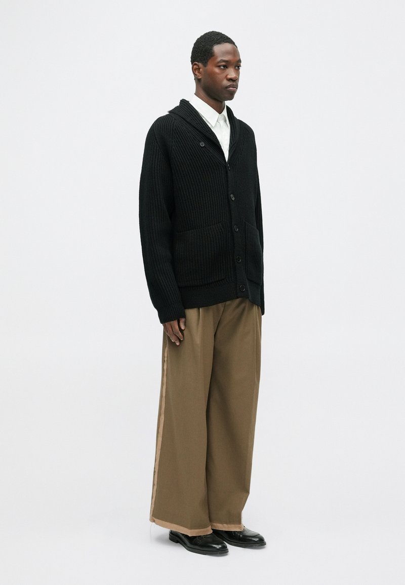 Cardigan nero a coste con chiusura a bottoni e due tasche frontali, abbinato a pantaloni larghi color sabbia e scarpe nere. Presenta un design con cappuccio.