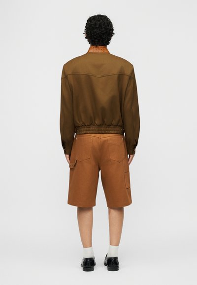 Άτομο με σγουρά μαλλιά στέκεται με την πλάτη γυρισμένη, φοράει καφέ μπουφάν τύπου bomber, καφέ σορτς που ταιριάζουν, λευκές κάλτσες και μαύρα παπούτσια σε ουδέτερο φόντο.