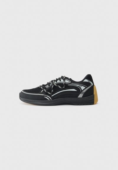 Zapatillas deportivas negras con parte superior de malla, suela de goma y acentos reflectantes. El diseño tiene superposiciones texturizadas y cordones en contraste.