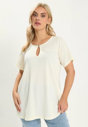 Blondynka ubrana w luźną kremową bluzkę z krótkim rękawem z małym wycięciem i serduszkowym guzikiem przy dekolcie, w połączeniu z niebieskimi dżinsami.