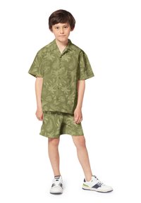 Olijfgroene overhemd met korte mouwen en bijpassende shorts met een subtiel bloemenpatroon, gecombineerd met witte en marineblauwe sneakers.